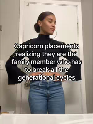 A woman stands in front of a white door, facing slightly to her right with a somewhat serious expression. She has dark hair pulled back, large hoop earrings, and is wearing a black long-sleeved top and jeans. Text overlayed describes a Capricorn's realization.