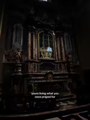 The image showcases a dimly lit, ornate church interior, with a focus on a high altar. The altar is richly decorated with gold accents, intricate carvings, and a central window. Two statues flanking the altar add to the religious iconography. A line of text is subtly placed near the bottom.