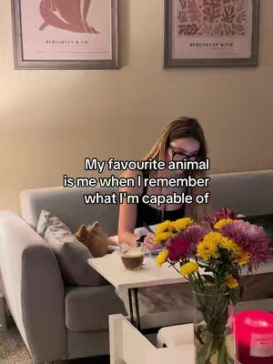 A woman with long hair and glasses is sitting on a grey couch and working. There are some flowers and a cup on the table in front of her.