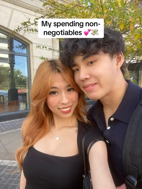 A selfie-style shot of a young couple smiling at the camera, taken outdoors. The woman, with long auburn hair, is on the left, with a necklace. The man is on the right with short, dark hair. They are in front of a building with some foliage visible, possibly a park or urban setting.