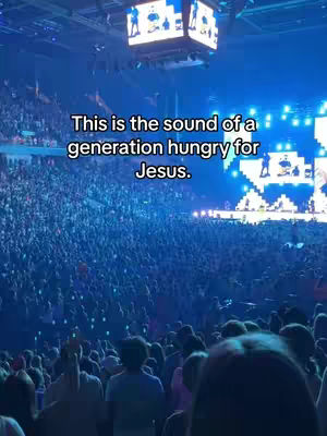 An image shows a vast crowd of people gathered in a large venue, presumably for a concert or religious event. The lighting is predominately blue. Projectors display images on the stage and large screens suspended above the audience.
