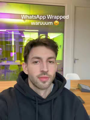 A man wearing a black hoodie is centered, looking at the camera against a background of an office with glass walls reflecting artificial lights.