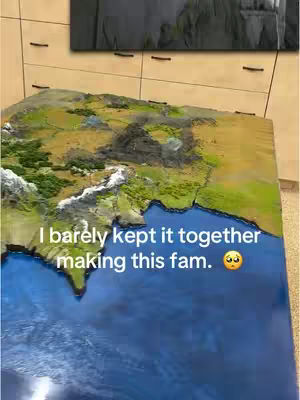A tabletop miniature model of a map, possibly depicting a fantasy world. The model shows landmasses with varying colors and textures, representing mountains, forests, and bodies of water. The background features a wooden cabinet and a glimpse of a framed picture.