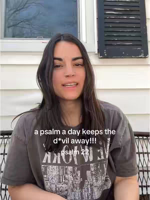 A woman with long dark hair is centered in the frame, smiling slightly. She's wearing a dark grey t-shirt with a graphic, seated in front of a white-paneled wall with a window and a dark shutter. Overlayed on the image are the words 'a psalm a day keeps the d*vil away!!!', followed by 'psalm 22'.