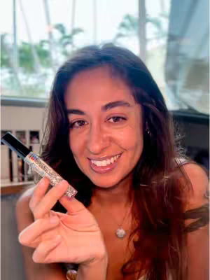 A woman with long brown hair is smiling and holding a lipstick tube towards the camera. She has a round pendant on her neck and appears to be in an indoor setting with a window in the background. The lipstick tube is silver and black. The overall setting is well-lit, suggesting a casual and inviting atmosphere.
