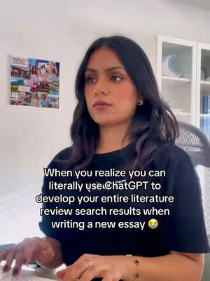 A medium shot shows a woman looking thoughtfully at the camera while sitting at a desk. She appears to be typing on a keyboard. The background includes a wall with a collage of images, a cabinet with items, and a window, suggesting a home office setting. Text overlay on the image states, 'When you realize you can literally use ChatGPT to develop your entire literature review search results when writing a new essay'.