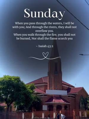 The image displays a church with a tall steeple against a gradient blue sky. Text is overlaid at the top, including the word 'Sunday' in a bold, white font. Beneath the word, a Bible verse from Isaiah 43:2 is presented in a clean, readable style. A heart-shaped graphic adds a decorative touch at the bottom of the text. The church's red-roofed architecture and its surroundings are clearly visible, giving the image a serene and inspirational mood.