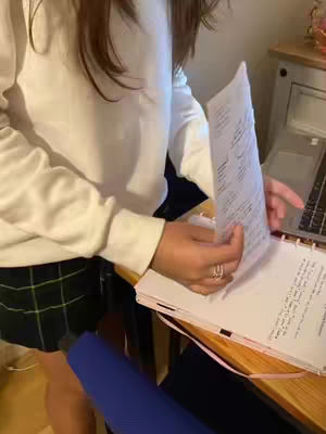 A person, wearing a white long-sleeved shirt and a plaid skirt, flips through pages of a notebook with lists while standing beside a wooden table. A laptop is partially visible in the background.