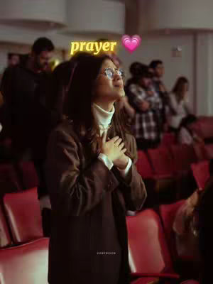 A woman with glasses and long hair is in a dark auditorium, her hands clasped over her chest as she looks upwards with an expression of devotion. The word "prayer" is styled in a yellow, glowing font above her head, coupled with a pink heart emoji.