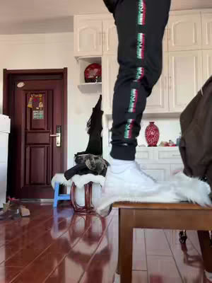 The image shows a person standing on a wooden surface, possibly a bench. The person's legs are visible, clad in black athletic pants with a colorful stripe. The person is wearing white sneakers, and the focus is on the shoes, which appear to have noticeable dirt marks. The background has clean white cabinets that could be in a residential interior.