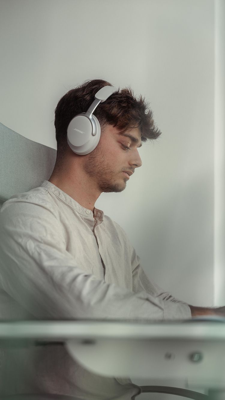 A man with short, dark hair is wearing gray Bose headphones, looking down and to the right. He is seated and wearing a light-colored, collared shirt. The background is a light gray color.