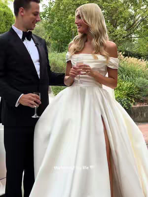 A couple is captured outdoors, the woman in a wedding dress and the man in a suit. They are holding glasses, likely celebrating. The background includes green foliage, suggesting a garden setting. The composition has a focus on the couple, highlighting their celebratory mood.