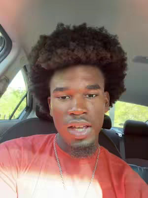 A man with an afro is sitting in the driver's seat of a car and looking directly at the camera. The background shows the car's interior, including the headrest of the back seat, and a glimpse of the outdoors through the window.