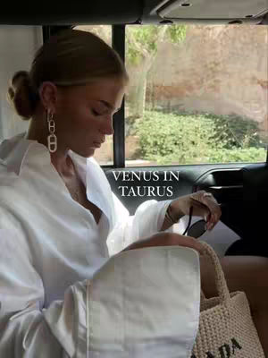 A woman wearing a white shirt and gold earrings looks down, sitting in a car next to a window with a view of green foliage and a stone wall. The text overlay "VENUS IN TAURUS" is centered on the image.