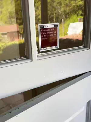 The image captures a close-up view of a white door with a window. Affixed to the window is a message displayed in a mock-up of a phone interface. The background visible through the window shows a green, natural environment.