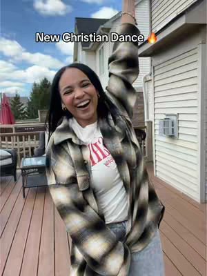 A woman with dark hair smiles broadly, raising her arm in a dancing pose. She wears a checkered shirt over a white t-shirt printed across the chest, with jeans. The background shows a wooden deck, a house with white siding.