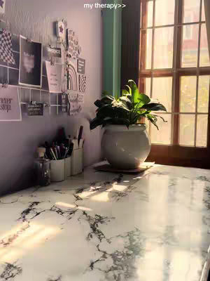A well-lit, aesthetically pleasing study space with a white and gray marble-patterned desk in the foreground. The composition features a window on the right, letting in natural light, and a wall on the left adorned with decorative items. The color palette is soft, with light purples, greens, and whites creating a calm atmosphere. A large potted plant sits on the desk, and the overall impression is one of a cozy, organized workspace.