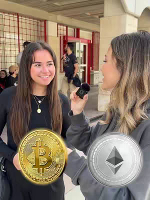 Two women stand outdoors, one holding a Bitcoin coin prop and the other an Ethereum coin prop, smiling at the camera. A man stands in the background, with a building and red-trimmed doorway behind them.