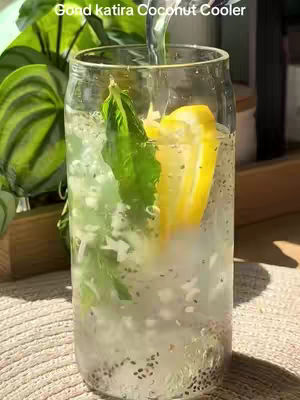 A glass of clear liquid with ice is being poured into it, the glass contains a slice of lemon and green leaves.