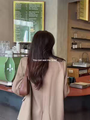 A medium shot of a woman from the back in a cafe setting. She is wearing a tan blazer and a brown purse. The background has a vintage coffee menu and some shelves.
