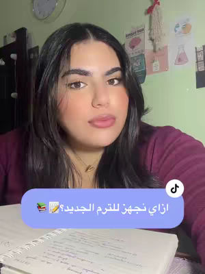 A young woman with long, dark hair and a burgundy top sits in a room with a partially visible clock on the wall behind her. She's looking directly at the camera with a neutral expression. Overlayed on the image is a rounded rectangle with text in Arabic asking about how to plan for the new semester, and a small TikTok logo in the top right corner. In the foreground, there is a notebook with handwritten notes.