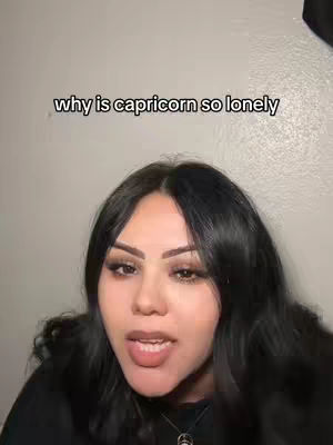 The cover image is a selfie-style shot featuring a woman. The woman is centered with her facial features prominently displayed. She has long, dark hair and is looking directly at the camera with a neutral expression. Her makeup includes eye shadow and lipstick, enhancing her features. Behind her, a plain, light-colored wall serves as the backdrop, keeping focus on her face. Overlaid on the image is the text "why is capricorn so lonely" in a simple, white font.