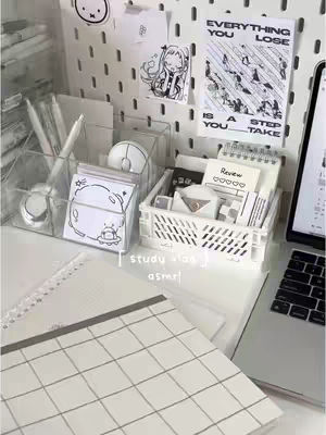 A well-lit desk setup with various stationery items and a laptop. The desk has a white pegboard with items pinned to it. A clear container holds pens, and there are sticky notes, a pen, and a mouse. A notebook and a laptop are also present. The overall aesthetic is clean and organized, with a focus on neutral tones and a minimalist style.