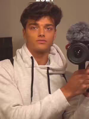 A young man with dark hair and a light gray hoodie is holding a camera with a microphone attached, appearing to film himself in front of a neutral background. The image has a slightly warm tone, suitable for personal vlogs.