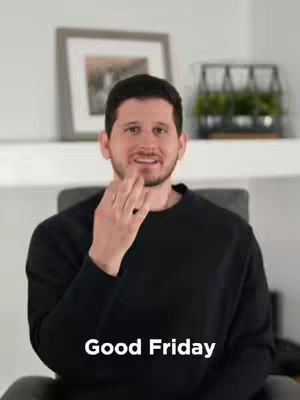 A man in a black sweater is signing ASL in front of a blurred background that has a framed picture hanging on the wall.
