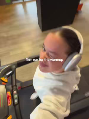 A woman is running on a treadmill in a gym setting. She wears a white hoodie and headphones. Text on the image reads "1km run for the warmup."