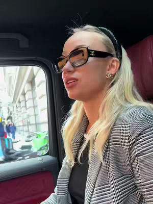 A blonde woman wearing a black and white checkered blazer and large sunglasses is sitting in the back of a car. The background includes a window with a blurry cityscape and a red car seat.