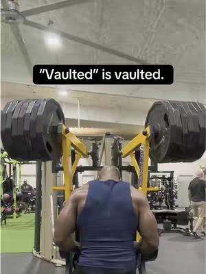 The image shows a muscular man from behind at a weightlifting gym. He is wearing a blue tank top. The man is positioned in front of heavy weight equipment with a sign that says "Vaulted" in a box on top of his head. The background includes various other gym equipment, several people, and a view of the ceiling.