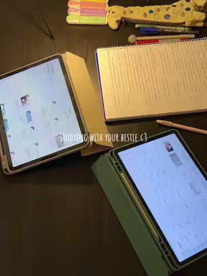 A top-down shot of a dark desk with two tablets, a spiral notebook, pens, a pencil, and colorful markers. The tablets have the screen on, and a notepad is open with handwriting. The overall vibe is organized and conducive to studying.