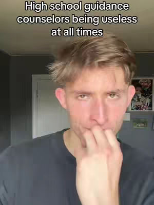 A young man with light skin and short blond hair is the central figure, positioned in front of a plain, light-colored wall and a partially visible door. He's wearing a black t-shirt, and his right hand is raised to his face, with his fingers touching his mouth, suggesting a pensive demeanor. The background is sparsely decorated, with a framed picture visible on the wall. The overall lighting is soft, giving the image a casual, indoor feel.