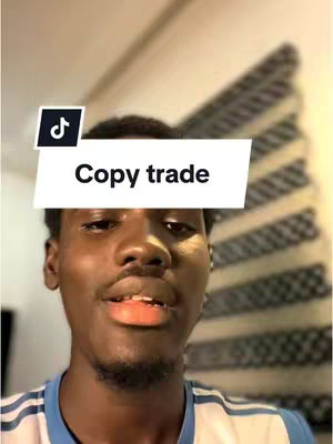 A young man of African descent is the central focus, with a short, neatly trimmed hairstyle and minimal facial hair. He's positioned in the foreground, looking towards the camera with a slight smile, his mouth slightly open. He wears a light blue and white striped shirt. Over his head is a white rectangle with the words "Copy trade" in a black font. The background is out of focus, featuring a patterned window blind and a corner of something else, but it is impossible to determine what they are.