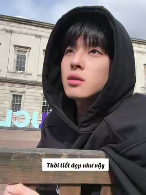 A young man wearing a black hooded sweatshirt sits outside with a building and sky visible in the background. A text overlay in Vietnamese is present. The man has short black hair and is looking upwards, with a thoughtful expression.