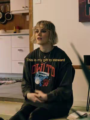 A woman with short blonde hair sits casually, looking slightly upwards. She wears a black sweatshirt with a graphic design. A text overlay reads 'This is my gift to steward'. The background is a domestic setting with cabinets and other furniture.