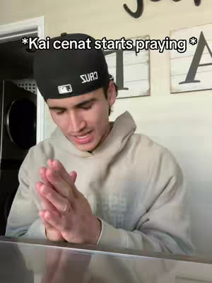 A young man is shown from the chest up, dressed in a hoodie and a baseball cap. He is positioned in front of a kitchen setup with a washing machine visible in the background. He is praying with hands pressed together. The lighting is soft, creating a slightly shadowed effect.