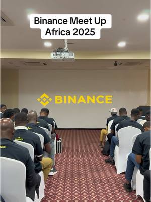 An indoor shot of a conference or meetup setting. A large banner with the Binance logo is at the back. Rows of people are seated facing the front with tables.