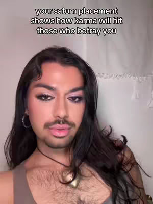 The image is a selfie-style shot featuring a person with dark hair and a mustache, wearing makeup. Text at the top says, 'your saturn placement shows how karma will hit those who betray you'. The subject is against a plain background.