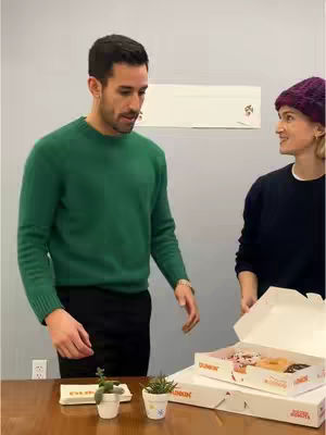 Two people in an office setting, a man in a green sweater is facing forward and looking down while a woman is standing next to him with a box of Dunkin donuts.