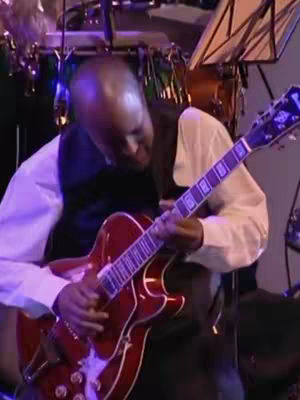 A man playing an electric guitar in a performance setting. He is wearing a white long-sleeved shirt with a dark vest over it. The guitar is red with white accents. In the background, there is a drum set and music stand.