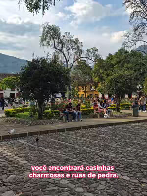 An outdoor shot of a public space, focusing on a cobblestone street in the foreground. In the mid-ground, there's a raised area with benches where several people are seated, appearing to be interacting or resting. Lush greenery, including bushes and trees, frames the seating area, creating a park-like setting. Behind the benches, there are buildings and additional trees, suggesting a town square or park setting. The sky is overcast with hints of sunlight, bringing a soft natural light that enhances the scene's warmth.