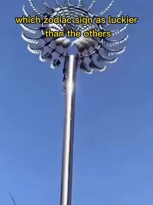 An upward shot of a sculpture against a bright blue sky. The sculpture is a tall, thin silver pole with a round, elaborate top that appears to be made of layered, circular elements. The top of the sculpture has a complex, almost floral-like design, with multiple reflective surfaces. Text is added on top of the image in yellow.