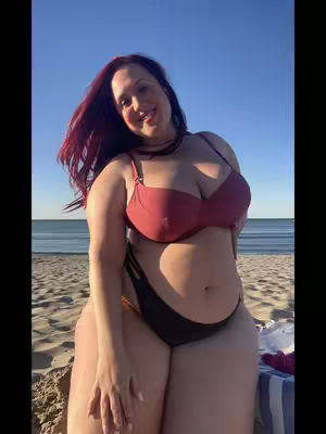 A woman in a red bikini top and black bikini bottoms stands on a sandy beach with the ocean and a clear blue sky in the background. She is smiling and looking towards the camera.