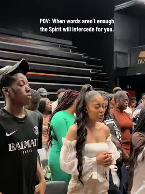 The cover image captures a diverse group of people, seemingly within a worship or religious setting. The foreground features a young Black man in a black shirt with white text and a black cap, and to his right, a young Black woman with her hair styled in a high ponytail, wearing a white top. The image is well-lit. Behind the two main subjects, several other people are visible, seemingly engaged in an activity that suggests prayer or some form of spiritual practice. The text overlay informs the context of the video.