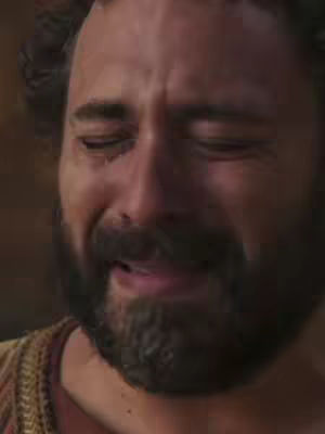 A close-up shot of a man with a trimmed beard, closed eyes, and tears streaming down his face. He wears a garment with a patterned shoulder, and the background is out of focus.