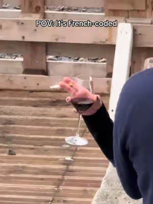 The image shows a person holding a glass of red wine and a lit cigarette, with the text 'POV: It's French-coded' overlaid. The scene appears to be outdoors, with wooden fencing in the background.