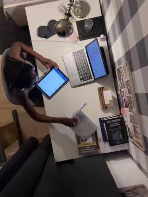 An overhead shot captures a woman sitting at a white table with a laptop and tablet. She's leaning over the table, holding a transparent document in her hand. Various items such as books, pens, a glass of water, and decorations are scattered on the table, with a striped wall in the background.
