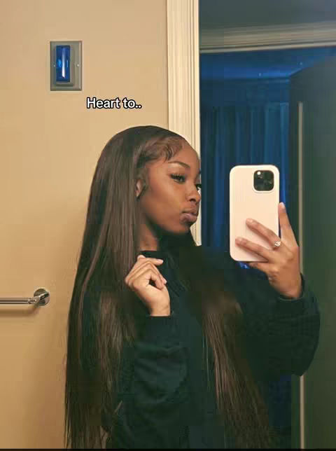 A selfie-style image taken in front of a light-colored bathroom wall. A young woman with long, dark hair is holding a phone up to capture the selfie, with the phone's screen not visible. Her gaze is directed towards the phone, and she appears to be looking at herself in the camera. There is a light switch visible on the wall.